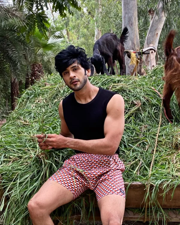 Man in a black vest and Whats Down maroon printed boxers sitting on hay beside goats smiling