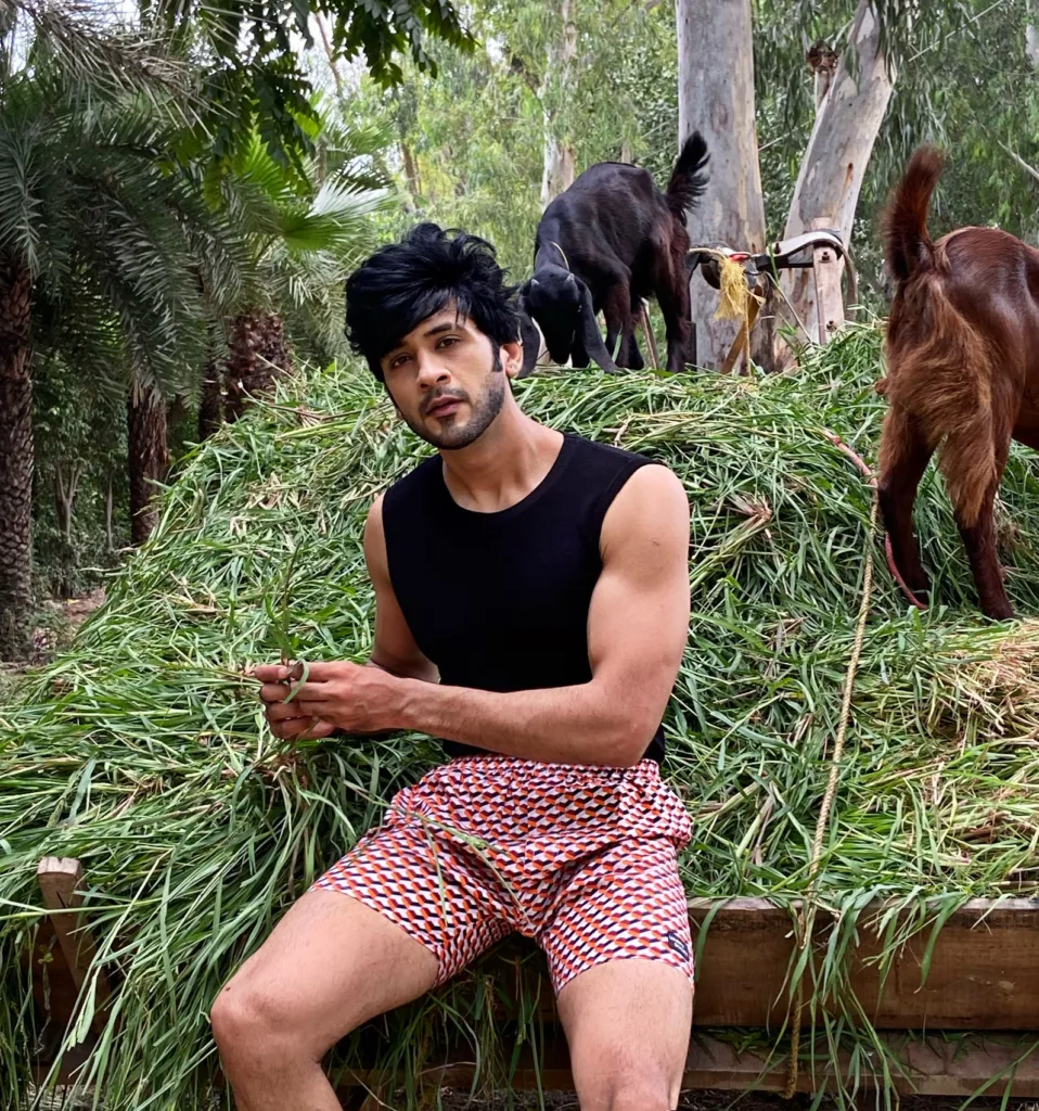 Man in a black vest and Whats Down maroon printed boxers sitting on hay beside goats smiling
