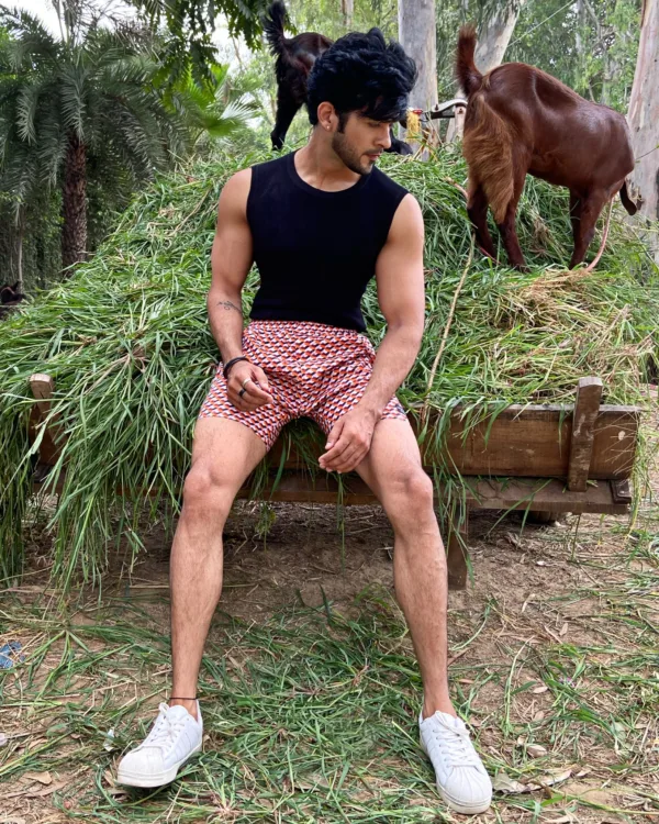 Man in a black vest and Whats Down maroon printed boxers sitting on hay beside goats