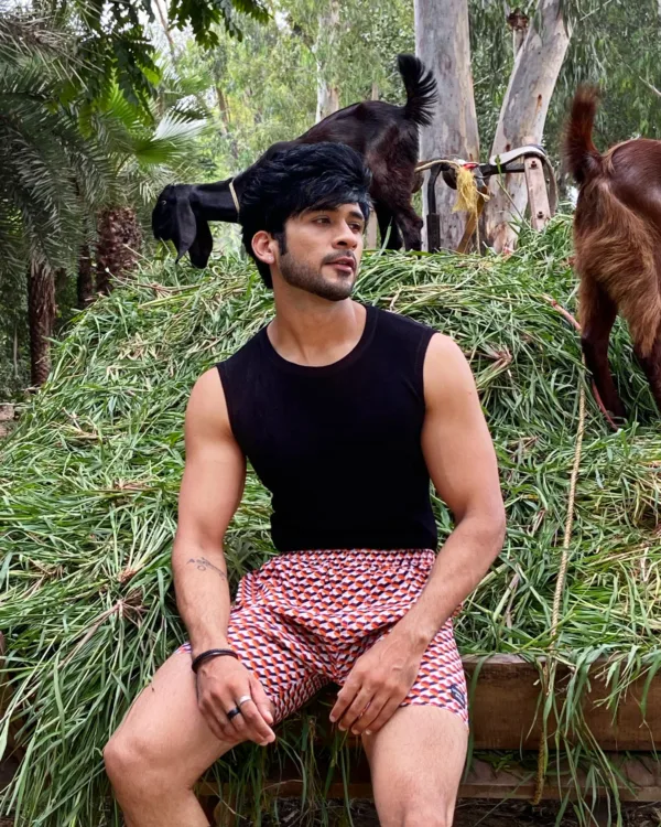 Man in a black vest and Whats Down maroon printed boxers sitting on hay with a goat