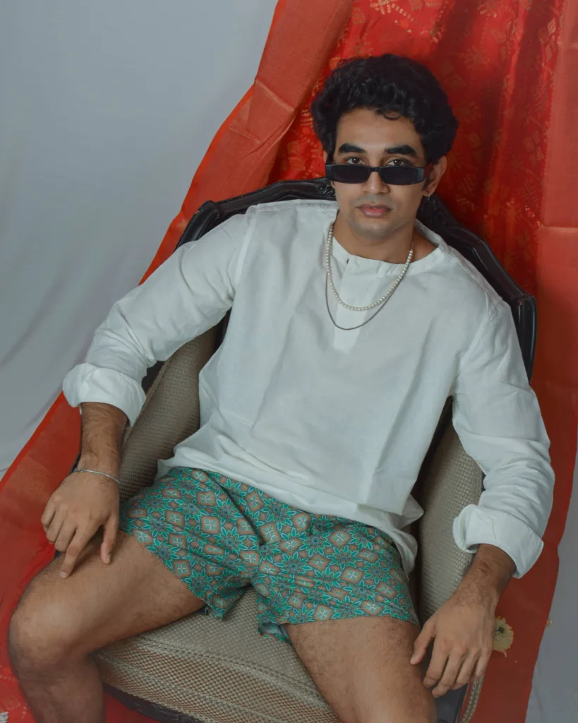 Man in a white sweatshirt, sunglasses and Whats Down green printed boxers on a red drape with marigolds on a chair