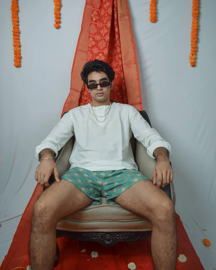 Man in a white sweatshirt, sunglasses and Whats Down green printed boxers on a red drape with marigolds seated, legs apart