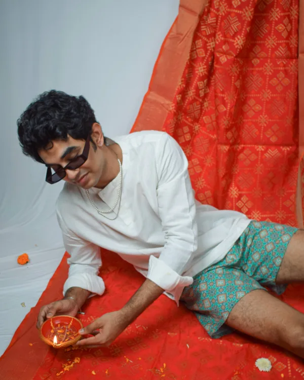 Man in a white sweatshirt, sunglasses and Whats Down green printed boxers on a red drape with marigolds holding a diya