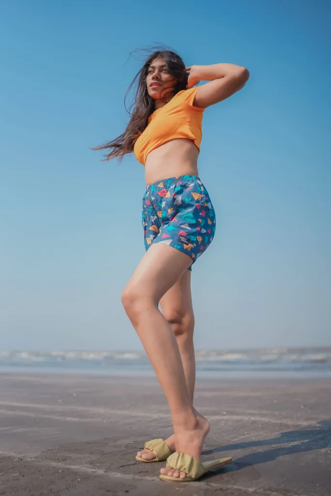 Woman in an orange crop top and Whats Down teal ice-cream printed boxers on the beach arms raised