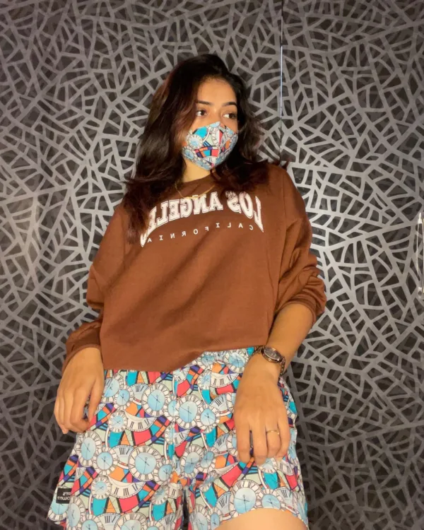 Woman in a brown sweatshirt and Whats Down multicolour printed boxers against a patterned wall wearing a printed face mask