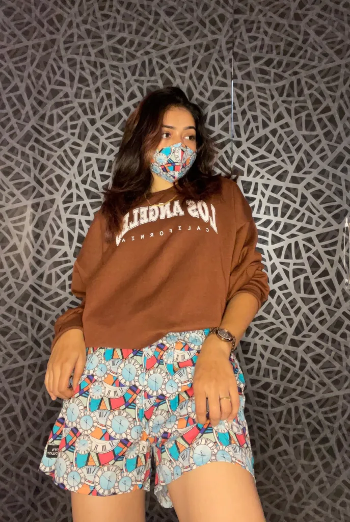 Woman in a brown sweatshirt and Whats Down multicolour printed boxers against a patterned wall wearing a printed face mask