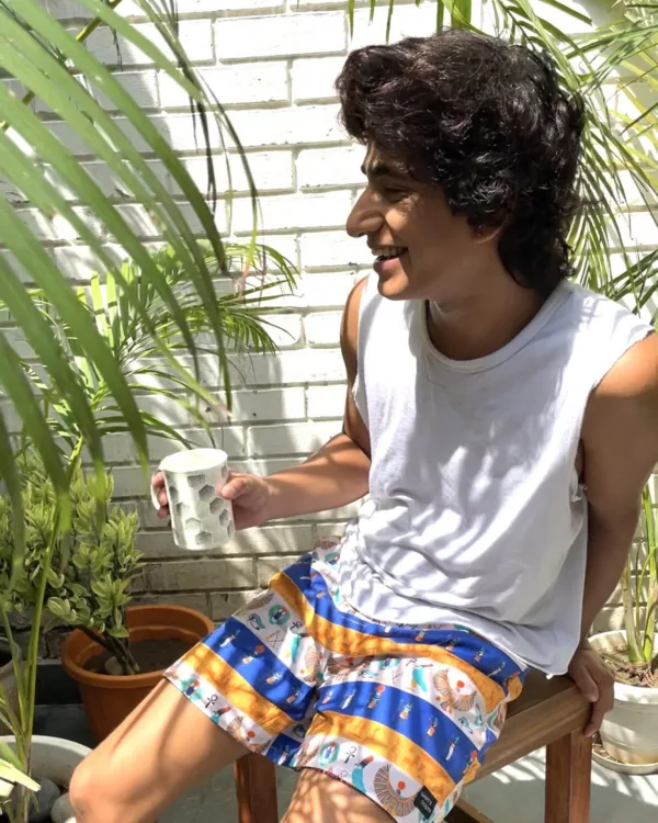 Man in a white vest and Whats Down blue-and-orange printed boxers on a chair by palm leaves laughing