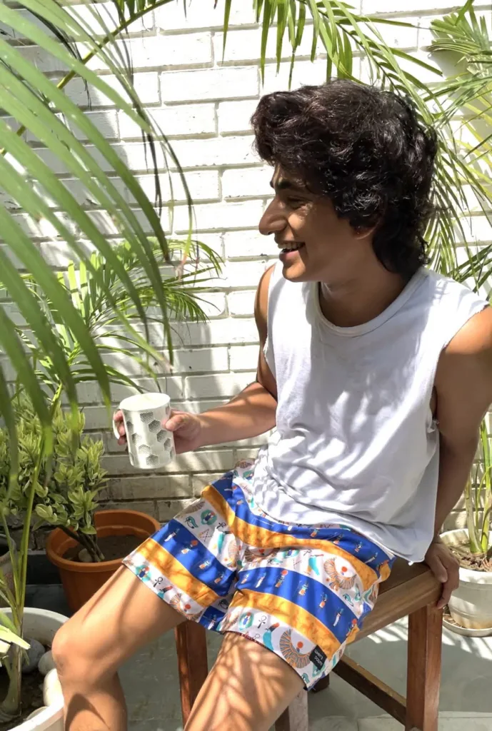 Man in a white vest and Whats Down blue-and-orange printed boxers on a chair by palm leaves laughing