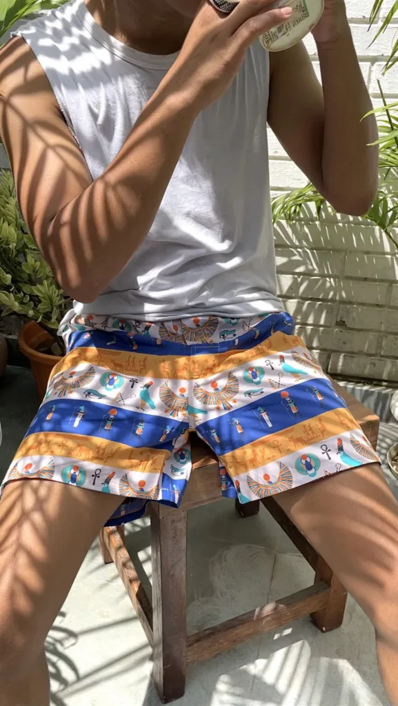Man in a white vest and Whats Down blue-and-orange printed boxers on a chair by palm leaves stretching
