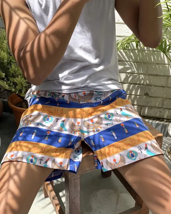 Man in a white vest and Whats Down blue-and-orange printed boxers on a chair by palm leaves stretching