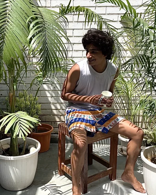 Man in a white vest and Whats Down blue-and-orange printed boxers on a chair by palm leaves sitting in the sun