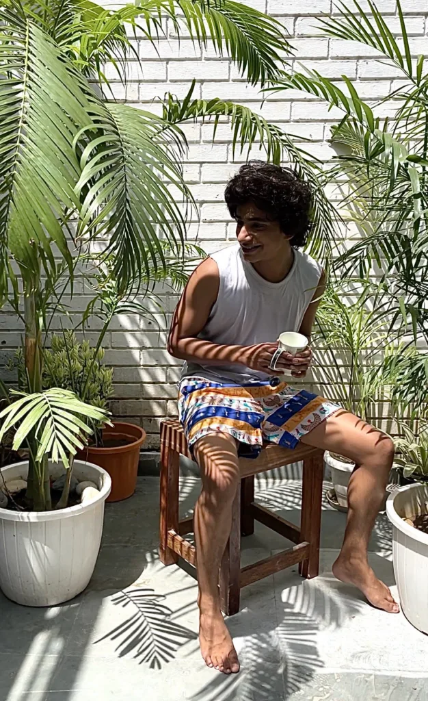 Man in a white vest and Whats Down blue-and-orange printed boxers on a chair by palm leaves sitting in the sun